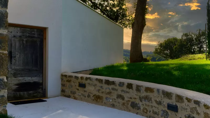 la porte d'entrée du gite la cubane en quercy rouergue ; la porte est en bois ancien, contrastant avec l'architecture résolument contemporaine ; sur la partie gauche de la photo, la pelouse, des arbres, le paysage valloné lointé éclairé par le soleil couchant