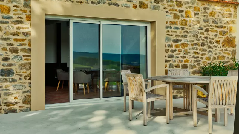 la terrasse du gite la cubane dans le tarn et garonne ; une table en bois, des fauteuils de jardin en bois autour, en arrière plan, la façade principale du gîte en pierre apparente, une baie vitrée permettant d'apercevoir la salle à manger intérieure