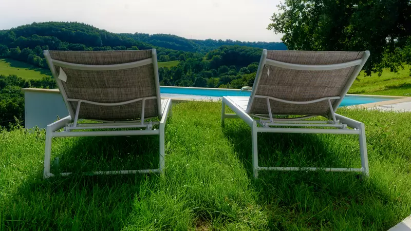 deux transats vus de dos sur la pelouse du gite la cubane en quercy rouergue ; au fond, la piscine, puis les vallons des gorges de l'aveyron, boisées avec quelques prés