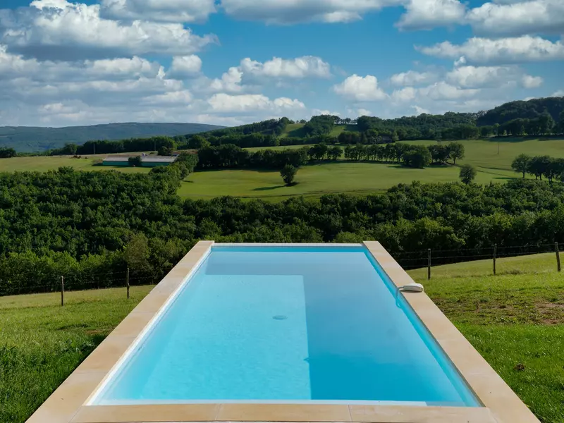 la piscine du gite de la cubane, vue en perspective au premier plan, avec son eau bleu turquoise ; en arrière plan, les côteaux du quercy rouergue avec prés et bois