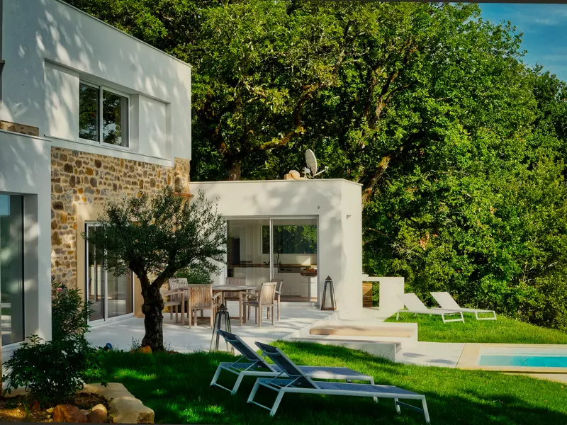 la façade sud du gite la cubane dans le tarn et garonne ; architecture contemporaine avec le bloc central sur deux niveaux en pierre apparente et enduit, le bloc cuisine sur un niveau en enduit blanc ; sur la terrasse, une table et des chaises ; sur le devant, deux transat et un olivier