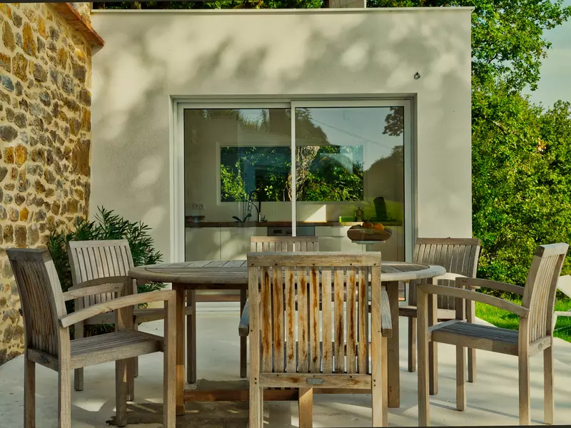 le coin repas extérieur du gite la cubane près de saint antonin noble val avec une table en bois, six fauteuils en boi ; dans le fond la cuisine, très contemporaine, accédant directement sur la terrasse par une large baie vitrée