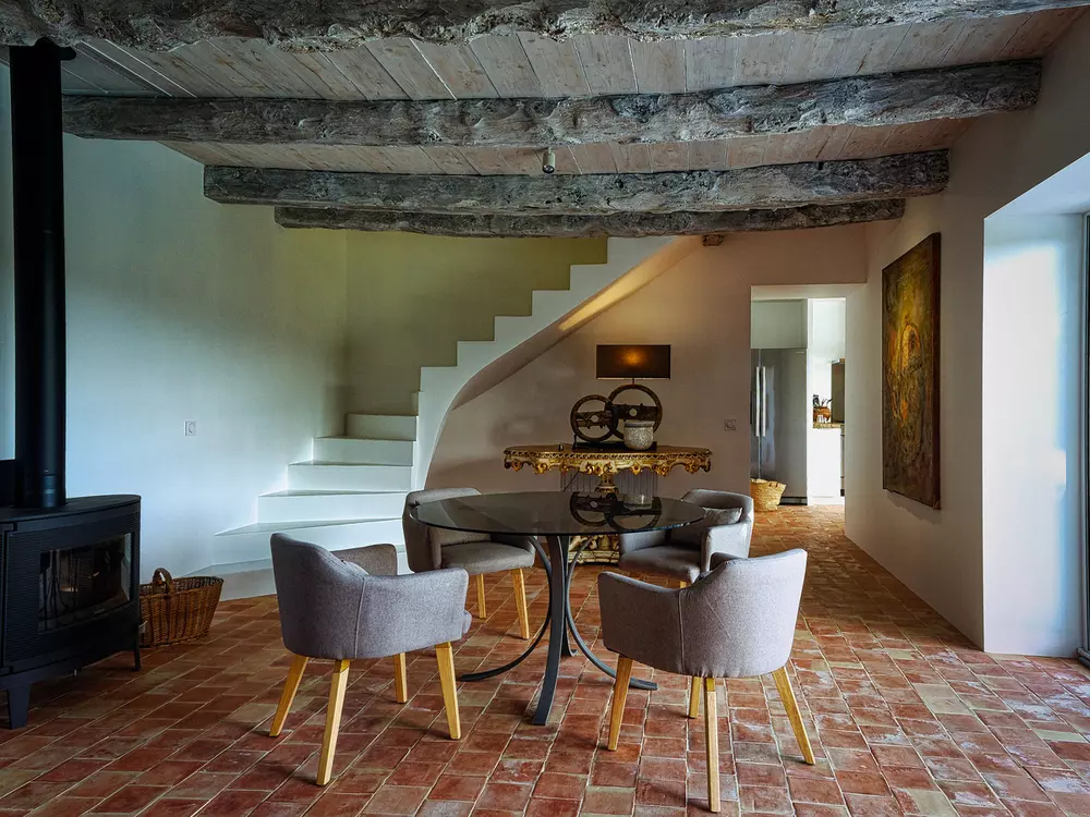 la salle à manger du gite la cubane dans le tarn et garonne ; une table en verre, quatre fauteuils, dans le fond une table-console de style baroque italien et la coursive qui mène à la cuisine ; sur le côté, un poële