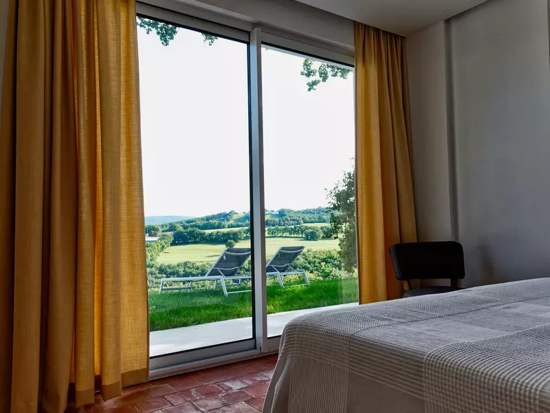la vue sur l'extérieur depuis la chambre du rez de chaussée du gite de la cubane en occitanie ; au premier plan, le coin du lit, puis deux transat et au loin les vallons et les prés