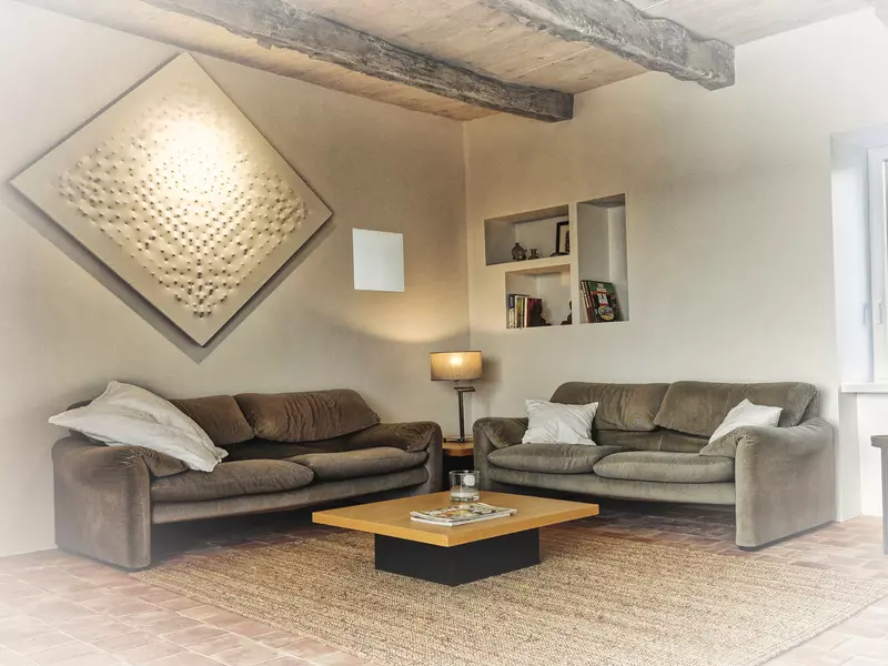 le coin salon du gite de la cubane dans le quercy, gorges de l'aveyron ; deux canapés, des coussins, une table basse avec une revue dessus ; sur le mur de gauche, un tableau contemporain, dans le fond trois niches avec livres et bibelots ; un fenestron donne un peu de lumière