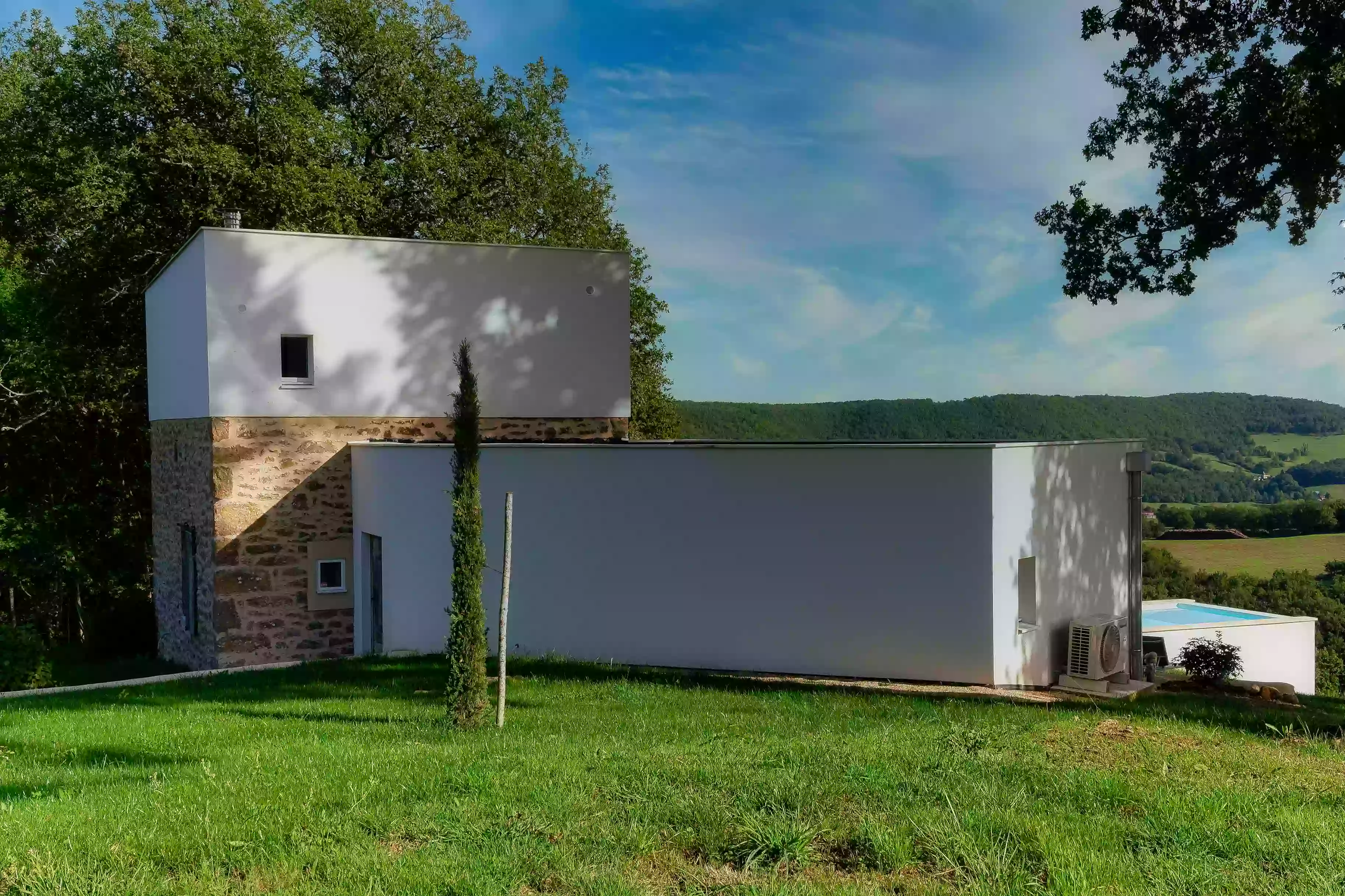 le gîte de la cubane, façade nord avec l'aile ouest à un niveau et la partie centrale sur deux niveaux, l'un en pierre apparente, l'autre enduit en blanc ; devant, la pelouse ; au fond, la piscine et dans le lointain les collines de Verfeil sur Seye