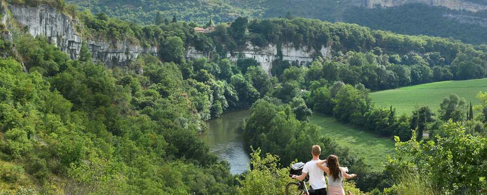 un couple faisant une promenade en vélo domine la vallée de l'aveyron à proximité de saint antonin noble val. En contrebas, la rivière. On aperçoit un hameau perché sur la falaise à gauche