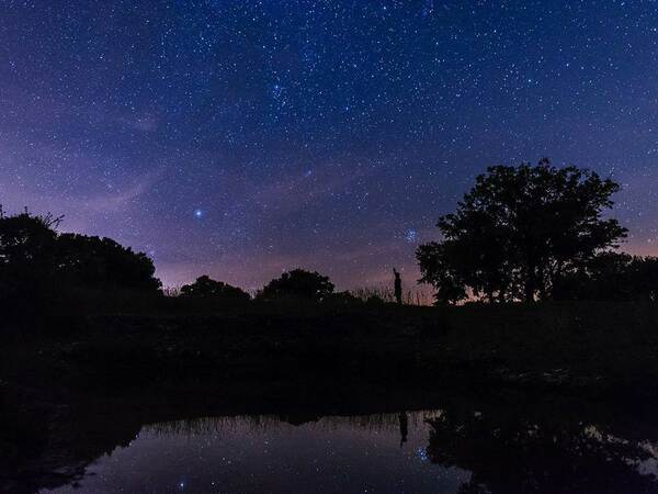 un ciel étoilé dans le triangle noir du quercy, département du lot en occitanie ; en premier plan, une étendue d'eau, puis dans le fond la silouhette d'un astronome et un arbre ; vers le haut, le ciel très pur, très clair