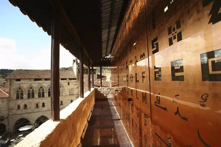vue de la coursive du musée champollion avec un mur recouvert de hiéroglyphes stylisés. Dans le fond, l'abbatiale de figeac et la terrasse d'un café sur une place