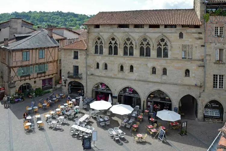 la grand placd de figeac, avec ses maisons en piere ou en brique et colombage et ses portes fenêtres de style gothique. Sur la plce un café avec ses tables, ses chaises et ses parasols