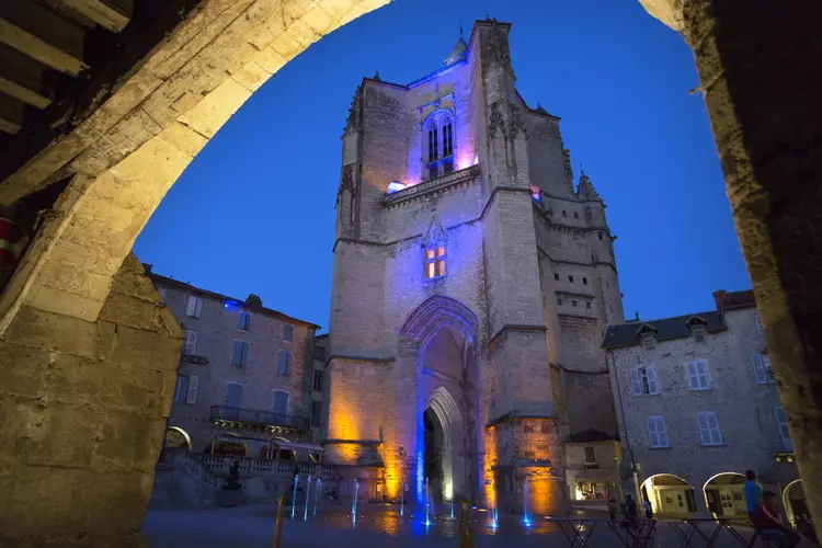 la place notre dame de villefranche de rouergue en aveyron, occitanie, illuminée la nuit : au travers d'une arcature, on voit l'abbatiale et sur la place la fontaine contemporaine